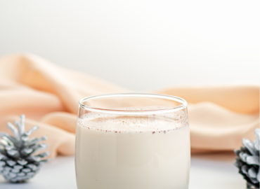 A short glass of white eggnog with a peach colored layer of fabric behind it and a pinecone on either side.