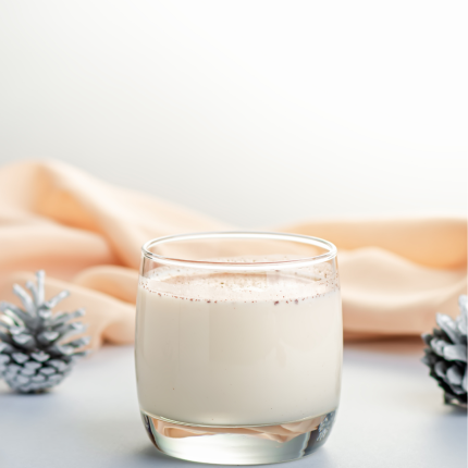 A short glass of white eggnog with a peach colored layer of fabric behind it and a pinecone on either side.