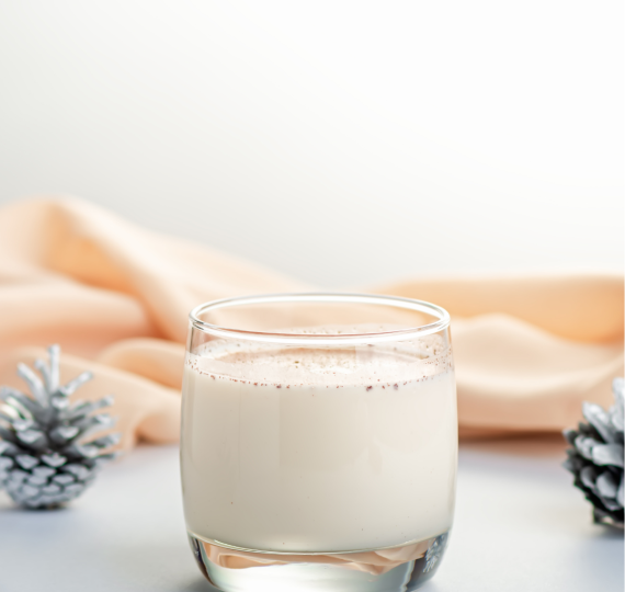 A short glass of white eggnog with a peach colored layer of fabric behind it and a pinecone on either side.